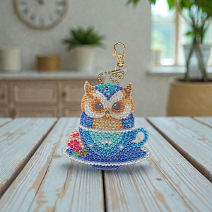 Puede incluir: Un llavero decorativo de búho, elaborado con brillantes pedrería en tonos azules, naranjas y blancos. El búho está posado en una taza de té, también adornada con pedrería. Se adjunta una cadena y un cierre dorados. Un accesorio encantador.