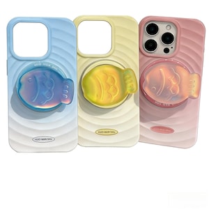 Puede incluir: Tres fundas para teléfono en colores pastel: azul claro, amarillo y rosa. Cada funda presenta un agarre emergente en forma de pez con tonos iridiscentes o amarillos. Las fundas tienen una textura ondulada y el texto "HUO MIN SHU".