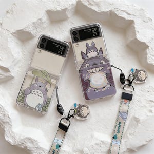 May include: Two clear phone cases with cartoon character designs. One case features a large gray character holding a leaf, and the other has a smiling gray character with a smaller character on top. Each case has a matching lanyard with a charm.