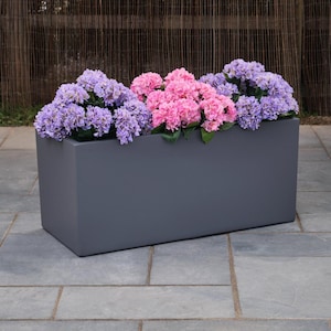 May include: A rectangular dark grey planter box filled with vibrant purple and pink hydrangea flowers. The container has a smooth, solid finish and a clean, linear design, providing a stark contrast to the lush, rounded clusters of petals.