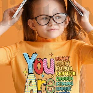 May include: A young person wearing a yellow turtleneck sweater with the words "You Are" in colorful, decorative lettering. The words "Brave, Smart, Helpful, Grateful, Enough, Strong, Unique, Loved" are also displayed. The phrase "DIGITALLY MADE YOURS" is at the bottom.