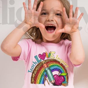 May include: A young person wearing a pink t-shirt with a colorful graphic. The graphic includes a rainbow, pencil, apple, and the words "Teach Love Inspire". The person is smiling and holding their hands up.