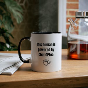 May include: White ceramic mug with a black interior and handle. The mug has the text "This human is powered by Chat GPTea" in black, and a teacup graphic. The mug is on a wooden surface.