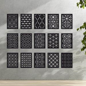 Può includere: Una collezione di quindici pannelli decorativi neri con intricati disegni geometrici ritagliati. I pannelli rettangolari sono montati su una parete grigio chiaro. I disegni variano, con stelle, cerchi e motivi floreali. Una pianta verde è visibile in alto a destra.