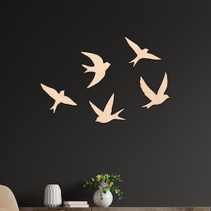 Puede incluir: Cinco golondrinas de madera en vuelo, dispuestas en un patrón circular, están montadas en una pared oscura. Los pájaros son de color beige claro y la pared es de un carbón profundo. La habitación también cuenta con un sillón beige, un mueble de madera y un pequeño otomán.