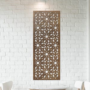 Op de afbeelding: Een rechthoekig, bruin houten wandpaneel met een geometrisch sterrenpatroon. Het paneel is bevestigd aan een witte bakstenen muur. De afbeelding toont ook een tafel en stoelen.