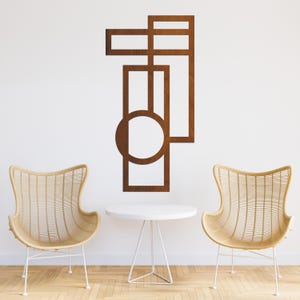 Puede incluir: Una pieza de arte mural geométrica marrón con recortes rectangulares y circulares. La obra de arte está montada en una pared blanca sobre una mesa blanca y dos sillas de mimbre. Las sillas son de color marrón claro.