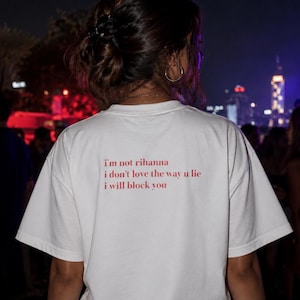 May include: White t-shirt with red text on the back: "I'm not rihanna, I don't love the way u lie, I will block you." The shirt is worn by a person at an outdoor event at night, with a city skyline in the background.