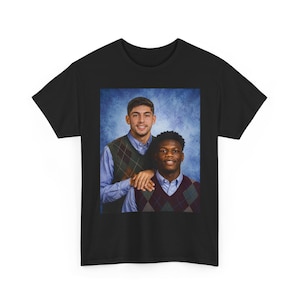 Può includere: Una t-shirt nera con una stampa grafica rettangolare di due persone in posa per un ritratto in stile scolastico. Entrambi indossano camicie azzurre sotto gilet a rombi su uno sfondo blu sfumato.