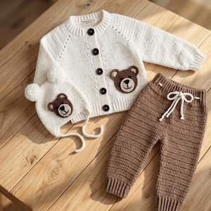 Puede incluir: Un conjunto de ropa de bebé de punto color crema que incluye un cárdigan, un gorro y un pantalón. El cárdigan tiene aplicaciones de oso marrón y botones. El gorro tiene orejeras y pompones. El pantalón marrón tiene un cordón.