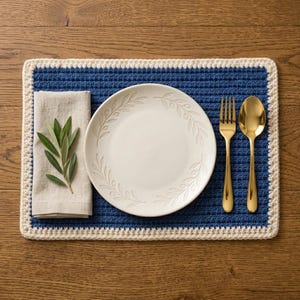 May include: A rectangular blue and cream crocheted placemat featuring a white ceramic plate with an embossed olive branch pattern, a folded beige linen napkin topped with a fresh green olive sprig, a gold fork, and a matching gold spoon.