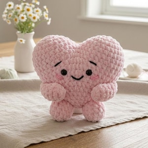 Op de afbeelding: Een handgemaakt, lichtroze gehaakt hartvormig pluche speelgoed met een lachend gezicht, zwarte ogen en roze wangen. Het pluche heeft kleine armen en benen. Madeliefjes in een witte vaas staan op de achtergrond.