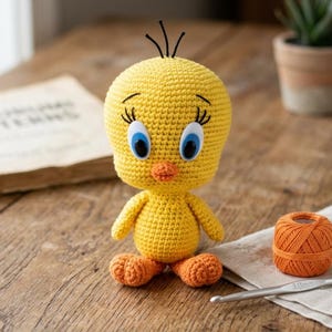May include: A handmade yellow crochet Tweety Bird doll with blue eyes and an orange beak and feet. The doll is sitting on a wooden surface next to a ball of orange yarn, a crochet hook, and a book.