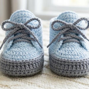 Può includere: Un paio di scarpe da ginnastica per bambini, lavorate all'uncinetto a mano in azzurro con dettagli grigi. Le suole e i lacci sono grigi e le scarpe sono legate con fiocchi. Il filato morbido e strutturato crea un aspetto accogliente.