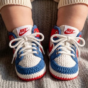 以下が含まれることがあります： ハイカットデザインのベビー用かぎ針編みスニーカー。つま先は白、アクセントは青、ディテールは赤で、タンにはナイキのロゴが入っています。白い靴紐が特徴です。