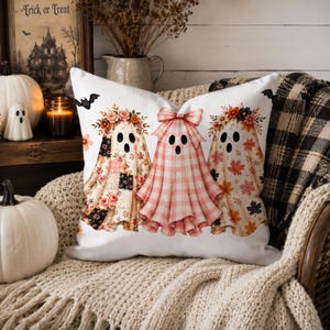 May include: A white square throw pillow featuring a graphic print of three ghosts. The left ghost has a floral pattern, the center ghost has a pink and white gingham pattern with a bow, and the right ghost has an autumn leaf pattern.