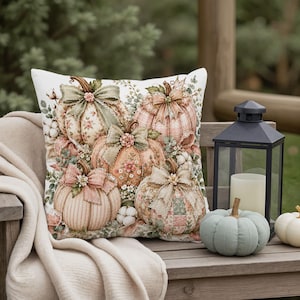 Peut inclure: Un coussin carré décoré d'un imprimé représentant quatre citrouilles ornées de nœuds en tissu, de motifs floraux et de textures à carreaux. Les citrouilles sont entourées de feuillage vert et de touches de coton blanc sur fond blanc.