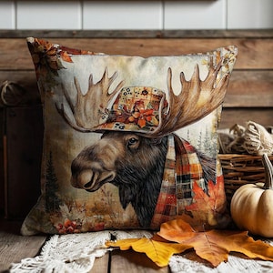 May include: A square throw cushion featuring a printed illustration of a moose wearing a plaid hat and scarf. The design includes autumn-themed colours, maple leaves, and a forest background. The cushion is displayed with a pumpkin and fall foliage.