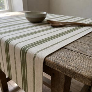 Peut inclure: Un chemin de table avec un motif de rayures verticales vert olive sur une base en tissu crème. Le chemin de table est posé sur une surface en bois avec un bol en céramique et deux cuillères en bois posés sur le matériau textile.