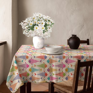 May include: A beige tablecloth with a repeating pattern of colorful fish in shades of pink, orange, green, and blue. A white vase with white flowers sits on the table, along with a white bowl and plate. A dark brown vase is also on the table.