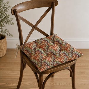 Puede incluir: Un cojín de asiento cuadrado con un patrón botánico en tonos naranja óxido, verde y crema, con detalles capitoné y lazos de tela de color claro en las esquinas, colocado sobre una silla de madera con respaldo en cruz y travesaño curvo.