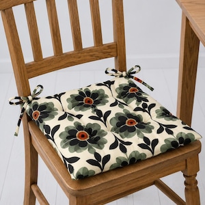 May include: A floral chair cushion with a cream background, featuring large black and green flowers with orange centres. The cushion is tied to a wooden chair with matching fabric ties. The chair is made of light brown wood.