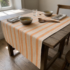 May include: A rectangular table runner featuring a pattern of vertical orange and cream-coloured stripes. The runner is draped over a wooden table set with two ceramic bowls containing soup, two wooden spoons, two folded napkins, and a clear glass.