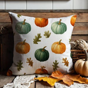 May include: A square throw cushion featuring a white background with a repeating pattern of orange and sage green pumpkins interspersed with small green and brown oak leaves. The fabric has a visible texture and a clean, finished edge design.