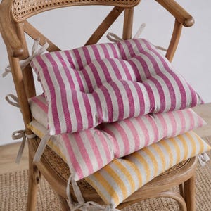 Puede incluir: Tres cojines de asiento a rayas en tonos rosa y amarillo apilados en una silla de madera. Los cojines, con rayas verticales, están sujetos a la silla con cintas neutras. La silla tiene un respaldo tejido.