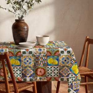 Puede incluir: Un mantel colorido con un diseño de patchwork que presenta limones, flores y patrones geométricos. La mesa está puesta con platos blancos, un cuenco y un jarrón con flores blancas. Sillas de madera rodean la mesa, creando un ambiente cálido y acogedor.
