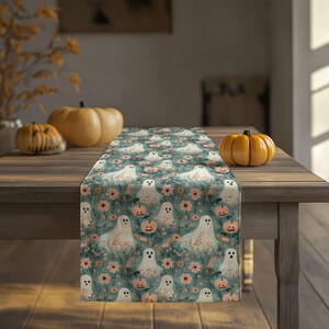 Op de afbeelding: Een rechthoekige tafelloper met een herhalend patroon van witte geesten, kleine oranje pompoenen en roze bloemen tegen een gedempte blauwgroene achtergrond. De stof toont een dichte print van deze seizoensgebonden motieven.