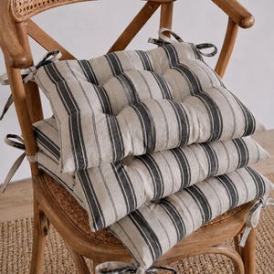 May include: A stack of three square seat cushions featuring a beige and charcoal grey vertical striped pattern. Each cushion is tufted with fabric ties at the corners for securing to a chair, made from a textured fabric with visible weave.