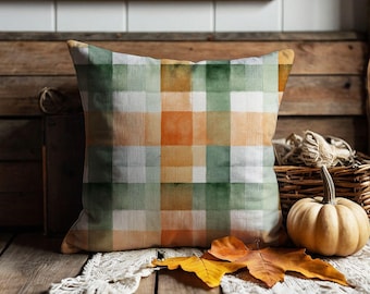 May include: A square throw pillow featuring a watercolor-style plaid pattern in shades of orange, forest green, and white. The fabric has a soft, blended appearance with intersecting vertical and horizontal stripes forming a grid across the surface.