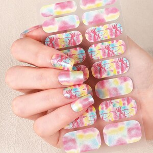 May include: A sheet of adhesive nail wraps featuring a tie-dye pattern in shades of pink, yellow, and blue, with some sections displaying a tropical palm tree silhouette design. The wraps are applied to fingernails in a gradient color scheme.