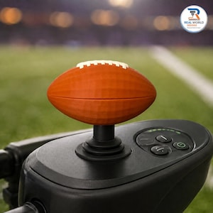 Puede incluir: Un mango de joystick con forma de balón de fútbol americano marrón con cordones blancos, montado sobre una base de control negra con botones circulares y un indicador de encendido. El mango tiene una superficie texturizada.