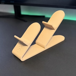 May include: A beige 3D printed controller stand with a dual-cradle design featuring rounded upright supports and a small embossed Xbox logo on the front base. The stand has a smooth, matte finish and a continuous, angular structure for holding devices.