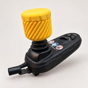 May include: A black electronic joystick controller featuring a bright yellow, cylindrical, diagonally ribbed handle. The base includes a circular dial, a green power button, a red stop button, and three circular safety warning icons printed on top.