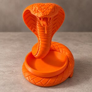 May include: A bright orange cobra snake figurine with a raised hood and open mouth, featuring a coiled body that forms a circular tray or dish base. The surface of the snake displays a detailed, textured scale pattern across its entire form.