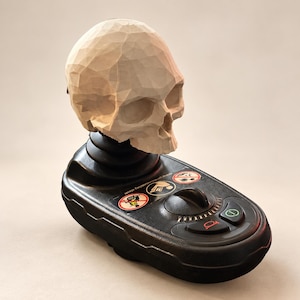 May include: A light-colored, faceted geometric skull sculpture mounted atop a black electronic wheelchair joystick controller. The controller features a circular speed dial, a horn button, a power button, and three circular safety warning icons.