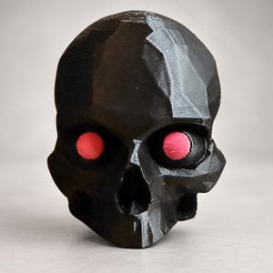 May include: A black, faceted skull sculpture featuring a geometric, low-poly design. The skull has two circular, bright pink eyes set deep within the eye sockets. The surface of the object displays visible horizontal 3D printed layer lines.