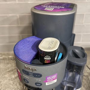 May include: A grey Ninja Thirsti drink system with a purple accent lid and a Peach Mango energy flavor pod inserted. The control panel features buttons for flavor, sparkling, and size, with a sticker reading Don't Grab Your Drink Until You Hear The Chime.