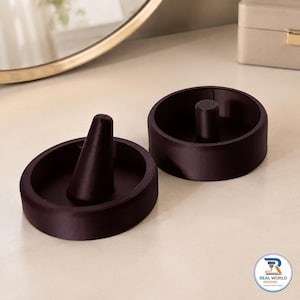 May include: Two dark brown circular ring holders with a smooth finish. Each holder features a central post for displaying rings. The left holder has a conical post, while the right holder has a cylindrical post. A logo reads Real World Designs.
