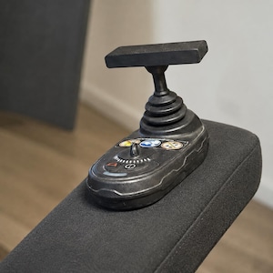 Puede incluir: Un mando tipo joystick negro con empuñadura en forma de T, fuelle de goma estriado y una base con tres iconos circulares que representan a una persona, un círculo azul y un círculo tachado, junto a un pequeño mando central y símbolo de encendido.