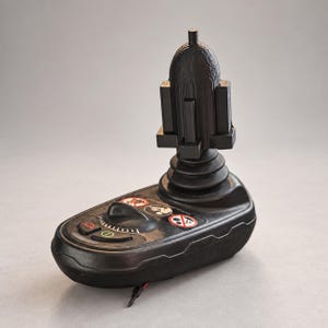 May include: A black joystick controller for a power wheelchair featuring a vertical handle with a textured top, a circular base with a speed adjustment dial, and three circular warning icons printed on the surface near the control buttons.