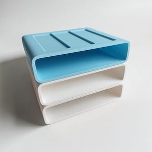 May include: A stack of three rectangular silicone soap dishes with rounded corners. The top dish is light blue with three drainage ridges on the surface, while the two dishes underneath are solid white. Each dish features a hollow center.