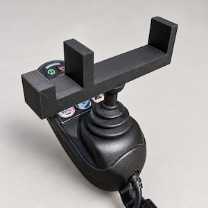 Puede incluir: Un soporte rectangular para teléfono impreso en 3D de color negro, acoplado a un mando tipo joystick negro. El mando cuenta con un fuelle de goma, una palanca central y un panel de control con iconos circulares, incluyendo uno de bocina.