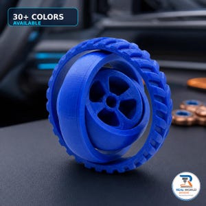 May include: A blue 3D printed fidget toy featuring a central wheel with five spokes encased within a rotating outer ring with a textured tread pattern. The image includes text stating 30+ COLORS AVAILABLE and a logo for REAL WORLD DESIGNS.