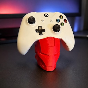 May include: A white wireless video game controller rests on a red, stylized helmet-shaped stand. The controller features two black joysticks, a black directional pad, and four colored action buttons labeled X, Y, A, and B.