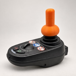 Puede incluir: Un controlador de joystick negro para silla de ruedas eléctrica con un mango cilíndrico de color naranja brillante. El panel de control incluye un botón de bocina, un botón de encendido, un dial de velocidad y tres iconos de advertencia.