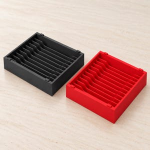 May include: Two square storage organisers, one in matte black and one in bright red. Each tray features a series of parallel vertical dividers creating multiple narrow slots for upright storage, with a low-profile frame and open top design.
